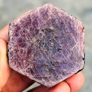 May include: A large, rough, purple ruby crystal with a hexagonal shape. The crystal has a natural, unpolished finish and shows striations and inclusions.