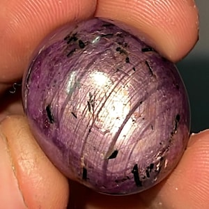 Könnte beinhalten: Ein glatter, ovaler, violetter Edelstein mit schwarzen Einschlüssen. Der Edelstein hat eine glänzende, polierte Oberfläche.