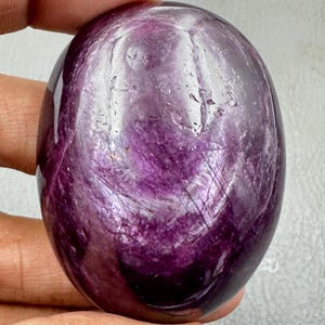 May include: A polished, oval-shaped gemstone with a deep purple and magenta hue. The surface displays natural internal inclusions, fine linear fractures, and a smooth, glossy finish that reflects light across its rounded, translucent structure.