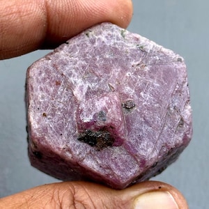 Könnte beinhalten: Ein rauer, sechseckiger Rubin-Edelstein mit einem tiefen, rötlich-rosa Farbton. Die Oberfläche hat ein strukturiertes Aussehen mit dunkleren Einschlüssen und einer kleinen, vorstehenden Kristallformation. Der Edelstein wird zwischen Fingern gehalten.
