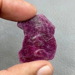 Op de afbeelding: Een ruwe, diep magenta robijn edelsteen. Het ongeslepen mineraal heeft een textuur met gebieden in lichtere, grijzige tinten. De robijn wordt tussen twee vingers gehouden tegen een neutrale achtergrond.