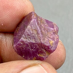 May include: A raw, uncut ruby gemstone held between fingers. The ruby is a deep, rich pinkish-red color with a rough, textured surface. The gemstone has a crystalline structure with visible facets and a natural, unpolished appearance.