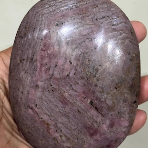 May include: An oval-shaped polished stone with a smooth, glossy surface. The stone features a mottled pattern of dusty rose, mauve, and deep purple hues, interspersed with dark grey and black mineral inclusions that create a subtle, layered texture.