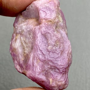 Peut inclure: Une pierre précieuse brute de rubis rose. La pierre naturelle présente une surface texturée avec des nuances de rose et une pointe de beige. Le rubis est tenu entre deux doigts sur un fond gris.