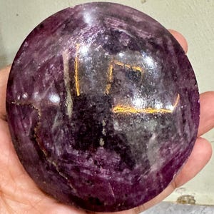 May include: A circular, polished stone cabochon featuring a deep purple and magenta color palette with natural veining and inclusions. The surface displays a smooth, reflective finish with visible internal textures and a distinct golden-yellow marking.