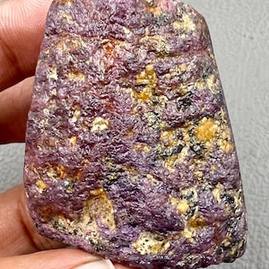 Puede incluir: Una gema de rubí en bruto y sin cortar, con un tono púrpura intenso, salpicada de áreas amarillas y negras. La superficie tiene un aspecto texturizado e irregular, mostrando la estructura cristalina natural del mineral. La gema se sostiene en una mano.