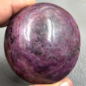 Könnte beinhalten: Ein polierter, kugelförmiger Edelstein mit einer tiefen Magenta- und Violettfärbung, durchzogen von sichtbaren dunklen Einschlüssen und natürlichen kristallinen Mustern, der eine glatte, abgerundete Textur und ein glänzendes Finish aufweist.