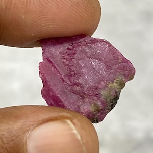 May include: A raw, uncut ruby gemstone in a deep pink colour. The natural stone has a textured surface and is held between two fingers. The ruby displays variations in colour and a slightly uneven shape.