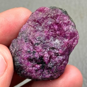 May include: A rough, uncut ruby gemstone with a deep magenta hue. The surface has a textured appearance with dark gray and black inclusions. The ruby is held in a hand, showcasing its natural, raw form. This gemstone is suitable for jewelry making.
