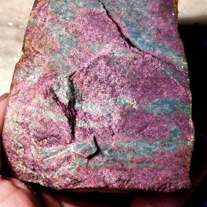 Puede incluir: Fragmento de roca en bruto con una llamativa paleta de colores. La piedra presenta una mezcla de magenta intenso, verde azulado y toques dorados. La superficie está texturizada con fisuras y variaciones naturales, mostrando la belleza en bruto del mineral.
