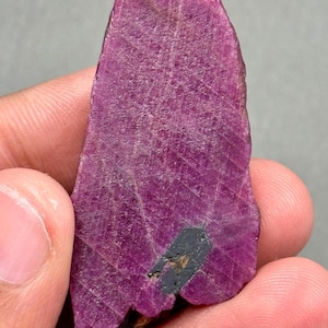 May include: A raw, natural ruby gemstone in a deep magenta colour. The stone has a textured surface with visible crystalline structures and a dark grey inclusion near the base. The ruby is held in a hand.