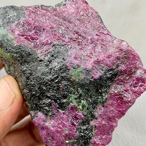 May include: A raw ruby specimen, displaying a striking contrast of deep pink and black. The natural stone features a textured surface with hints of green, revealing its crystalline structure.