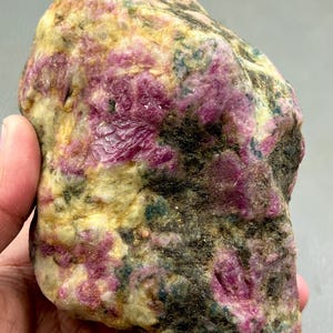 May include: A raw, unpolished mineral specimen featuring vibrant magenta and deep pink crystalline inclusions embedded within a textured, off-white and dark grey matrix. The stone displays a natural, irregular shape with varied surface patterns.