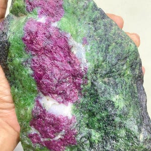 Op de afbeelding: Een ruwe robijn in zoisiet steen. De steen heeft een levendige kleurenmix, met diepe magenta robijninsluitsels tegen een groene zoisietbasis. De steen heeft een natuurlijke, onbewerkte uitstraling.