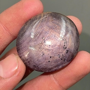 Peut inclure: Une pierre précieuse ronde de couleur violette avec une surface lisse et polie. La pierre a une qualité irisée unique avec des stries blanches et argentées.