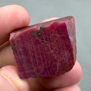 Può includere: Una gemma di rubino grezza e non tagliata, di colore rosso intenso. Il rubino ha una forma rettangolare con linee naturali visibili e alcune inclusioni verde scuro. La gemma è tenuta in una mano.