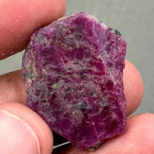 Pode incluir: Uma gema de rubi bruta, não lapidada, segurada entre os dedos. O rubi exibe uma cor vermelha profunda, com áreas de sombreamento mais escuro e uma superfície texturizada. A gema tem uma forma natural e irregular.
