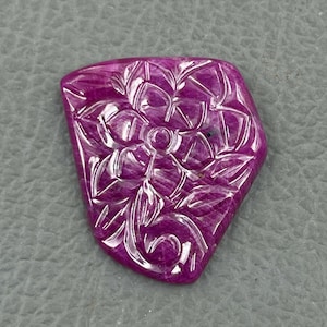 Op de afbeelding: Een gegraveerde roze edelsteen met een bloemmotief. De edelsteen is in een vrije vorm gesneden en heeft een gepolijste afwerking.
