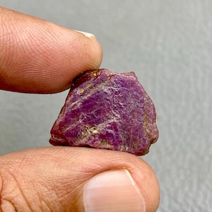 May include: A raw ruby gemstone, held between fingers. The ruby is a deep magenta colour with visible texture and golden inclusions. The gemstone is irregularly shaped, showcasing its natural, raw form.