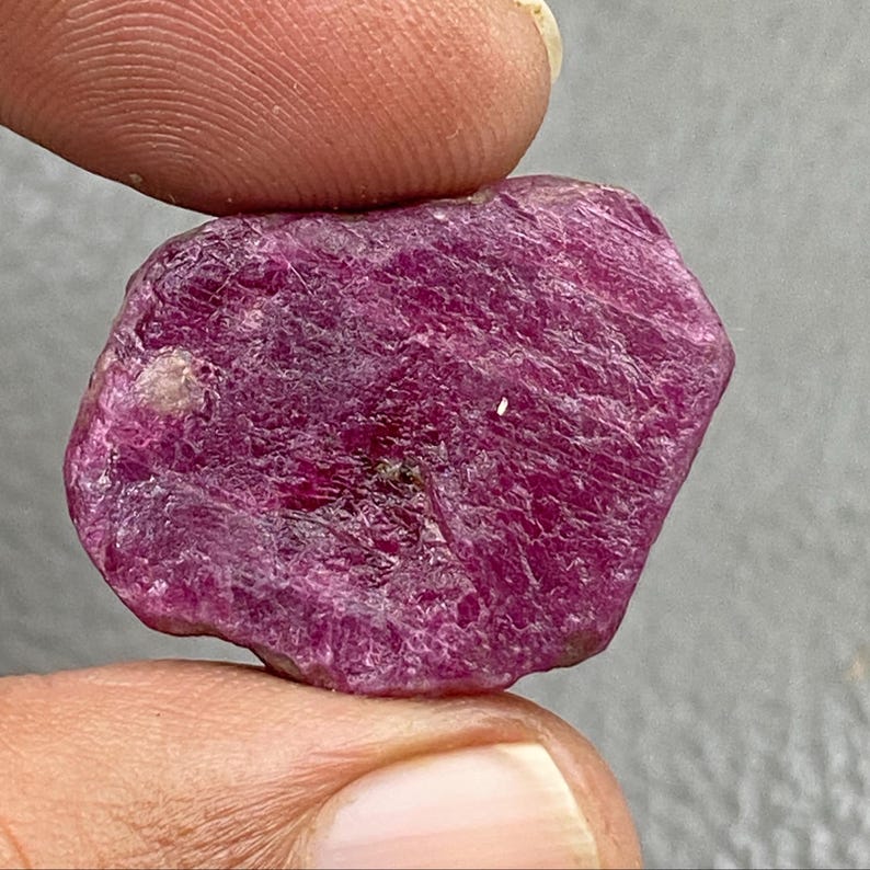 May include: A raw, uncut ruby gemstone. The ruby is a deep, saturated red-purple color with a rough, textured surface. The gemstone is held between two fingers, showcasing its natural form and color.