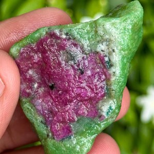 May include: A raw, uncut gemstone with a vibrant color palette. The stone features a striking combination of deep magenta and bright green hues. The rough texture and natural form of the stone are clearly visible, showcasing its organic beauty. The gemstone is held in a hand.