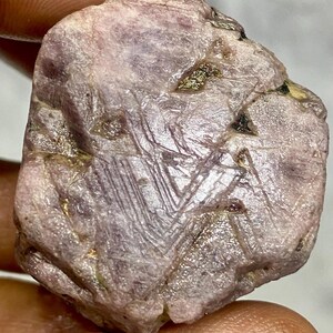 May include: A rough, uncut gemstone with a pale purple hue. The surface displays intricate linear patterns and small, dark inclusions. The stone has a natural, irregular shape and a slightly textured appearance, showcasing its raw, unpolished state.