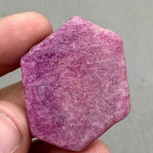 May include: A rough-cut, hexagonal ruby gemstone. The ruby is a deep pinkish-red colour with visible surface texture and natural lines. The gemstone is held between two fingers against a neutral background.