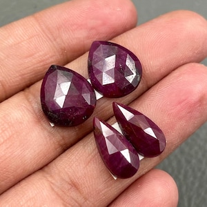 Puede incluir: Cuatro piedras preciosas de rubí rojo oscuro facetadas, en varias formas de lágrima. Las gemas se muestran sobre un fondo neutro, mostrando sus superficies pulidas y reflejos internos. Las piedras varían en tamaño y están talladas.