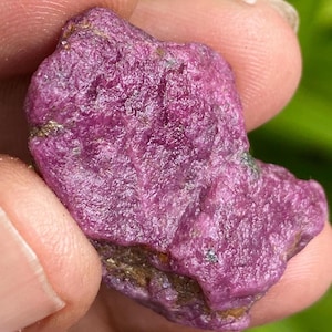 Può includere: Una gemma di rubino grezza e non tagliata, di colore magenta intenso. La superficie ha un aspetto strutturato e irregolare con sfumature di marrone e verde. La gemma è tenuta in una mano, mostrando la sua forma naturale e grezza.