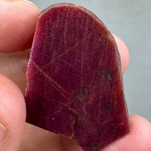 Può includere: Una gemma di rubino grezza, di forma irregolare. Il rubino è di un colore rosso intenso e ricco, con inclusioni e striature naturali visibili. La pietra è tenuta tra le dita, mostrando il suo stato naturale e non lucidato. Minerale naturale.