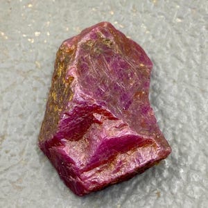Può includere: Una gemma di rubino grezza, non tagliata, di colore rosso-viola intenso. La pietra naturale ha una struttura cristallina con striature visibili e una superficie leggermente irregolare. Sono visibili sfumature dorate sui bordi.