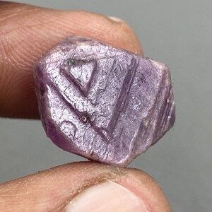 Può includere: Primo piano di una gemma viola, sfaccettata e dal taglio grezzo. La gemma ha una forma triangolare ed è posizionata su uno sfondo grigio.
