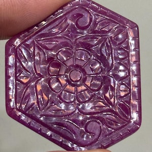 Peut inclure: Une pierre précieuse hexagonale de couleur magenta profond sculptée avec un motif floral central entouré de feuilles et de vignes. Les bords de la pierre présentent une série de petites encoches rectangulaires régulièrement espacées.
