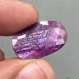 以下が含まれることがあります： 深紅色のファセット加工された楕円形の宝石。磨かれた表面には内部構造と光の反射が見えます。石は2本の指で持たれており、そのサイズと形状が示されています。