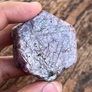 May include: A large, purple, faceted gemstone with a rough, natural texture. The gemstone has a hexagonal shape and is a raw, unpolished crystal.