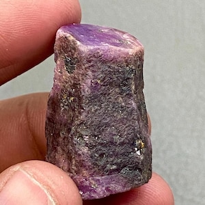 Może przedstawiać: Surowy, nieoszlifowany kamień szlachetny rubinu. Rubin ma głęboki fioletowy odcień z szorstką, teksturowaną powierzchnią. Kamień szlachetny jest trzymany między dwoma palcami, ukazując jego naturalny, nieoszlifowany stan. Kolor rubinu to bogaty, nasycony odcień fioletu.