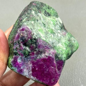 Könnte beinhalten: Ein roher Rubin in Zoisit-Edelstein. Der Stein weist eine lebendige Mischung aus Farben auf, darunter tiefes Magenta, Waldgrün und Schwarz. Die Oberfläche hat ein strukturiertes Aussehen, das die natürliche Bildung des Minerals zeigt.