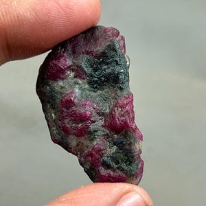 May include: A rough-cut ruby gemstone with a deep red hue and dark gray matrix. The ruby is held between fingers, showcasing its natural, unpolished texture. The gemstone has a raw, organic appearance.