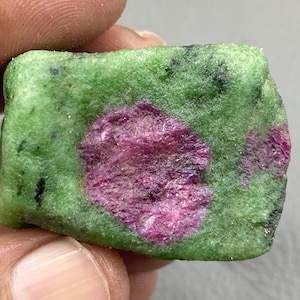 May include: A rough-cut gemstone featuring a vibrant green matrix with patches of deep pink and magenta. The stone has a natural, unpolished appearance, showcasing its raw mineral composition. Small black inclusions are scattered throughout.