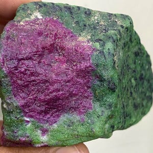 Può includere: Una gemma grezza con un vivace centro magenta circondato da una matrice verde macchiata. La superficie della pietra ha un aspetto strutturato, che mostra un'estetica naturale e non lucidata. La combinazione di colori crea un contrasto sorprendente.