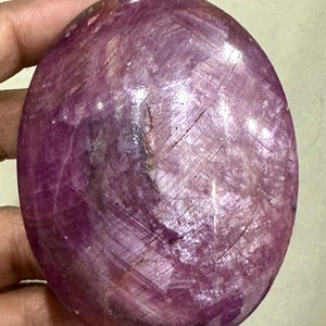 Könnte beinhalten: Ein oval geschliffener Rubin-Edelstein in tiefen Magenta- und Violetttönen mit sichtbaren natürlichen Einschlüssen, kristallinen Strukturen und einer glatten, reflektierenden Oberflächenbeschaffenheit über den gesamten Stein.
