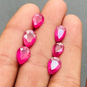 Op de afbeelding: Zes roze peervormige edelstenen, waarschijnlijk robijnen, worden op een hand getoond. De edelstenen zijn gefacetteerd en hebben een hoge glans.
