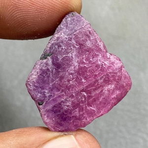 May include: A rough, uncut ruby gemstone. The ruby is a deep pinkish-red color with a slightly uneven surface. The gemstone is held between two fingers, showcasing its natural, raw form. The ruby has a small black inclusion.