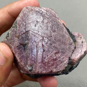 May include: A raw, uncut ruby gemstone with a deep pink colour. The surface displays natural lines and textures, with areas of darker, almost black, mineral deposits. The ruby is held in a hand, showcasing its raw, natural beauty.