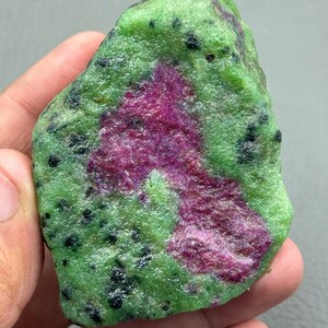 以下が含まれることがあります： 緑色のマトリックスに黒いインクルージョンが散りばめられた、未加工の宝石。中央部は、豊かな深紅の色合いを示しています。手で持たれており、自然で未研磨の質感が現れています。
