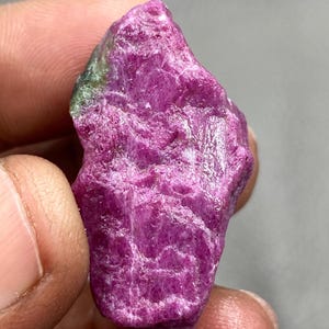 Può includere: Un cristallo di rubino rosa grezzo con inclusioni verdi. Il cristallo è lungo circa 5 cm e ha una forma irregolare e grezza.