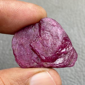 Peut inclure: Une pierre précieuse de rubis brute et non taillée, de couleur magenta foncé. La pierre naturelle présente une surface texturée et une forme irrégulière, tenue entre deux doigts. Le rubis est d'une teinte rouge-violet vibrante, révélant son état brut et non poli.