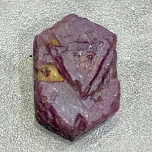 Pode incluir: Uma gema bruta, não cortada, de cor vermelho-púrpura, com uma estrutura cristalina. A pedra tem uma forma irregular com algumas manchas mais claras. A superfície tem uma qualidade ligeiramente refletora.