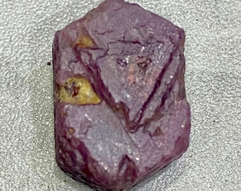Puede incluir: Una gema en bruto, sin cortar, de color rojo púrpura, con una estructura cristalina. La piedra tiene una forma irregular con algunas manchas más claras. La superficie tiene una calidad ligeramente reflectante.