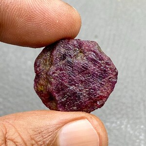 Peut inclure: Une pierre précieuse de rubis brute et non taillée, tenue entre les doigts. Le rubis est d'une couleur rouge profond avec une texture de surface visible et des inclusions naturelles. La pierre a une forme irrégulière et organique.
